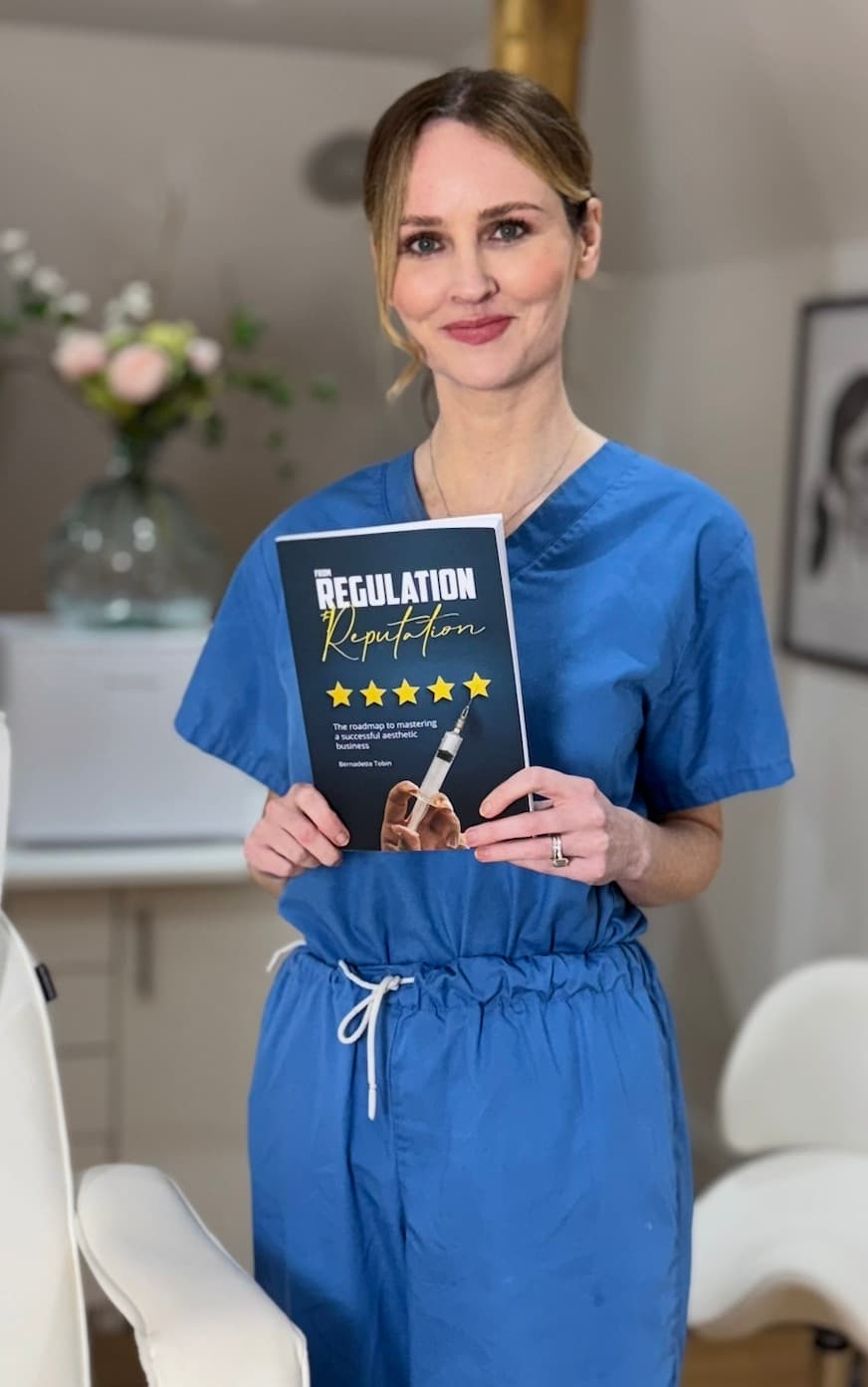 Bernadette Tobin RN MSc in her clinic, holding her book From Regulation to Reputation.