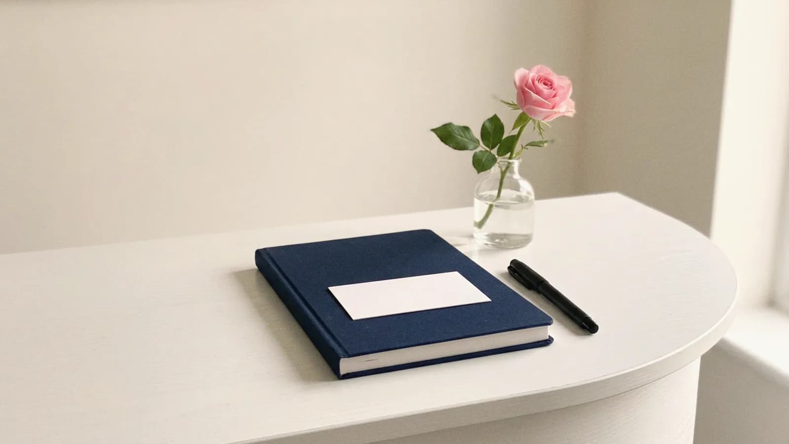 A clinic reception desk with a closed diary, a blank card and a pen beside a single pink rose.